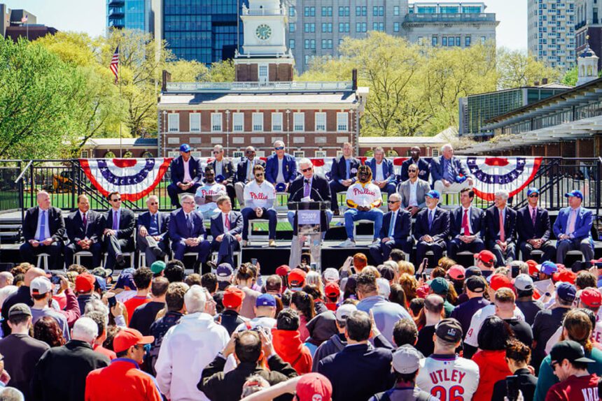 A crowd of people are gathered in front of a stage set up in front of Independence Hall. There are MLB players and officials on the stage making a major announcement, Philadelphia was chosen to host the MLB All-Star Game in 2026. Red, white, and blue flags are hung behind them/