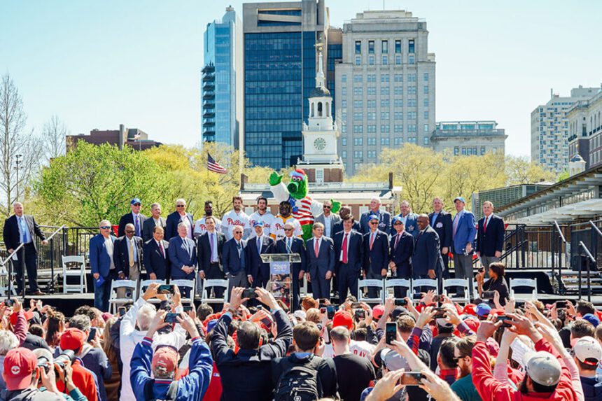 The Phillies Phanatic is shown in the center, behind two rows of men standing on a stage set up in front of Independence Hall. A crowd if people are shown below the stage, taking pictures with their phones. Many of them are wearing baseball caps. 