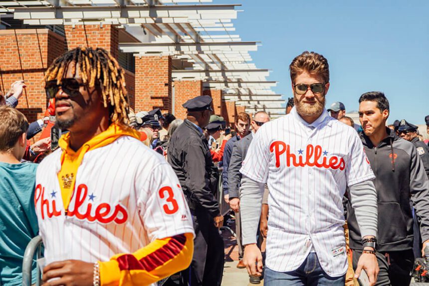 Phillies All-Stars Odubel Herrera and Bryce Harper are shown wearing their jerseys and walking past the camera. Crowds of people are behind them, gathered for a press conference by the Visitor Center.