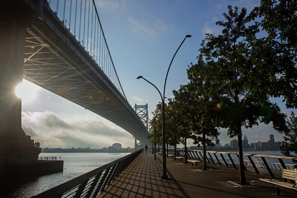 The bridge is shown off to the left, a walkway down the center is lined with benches and trees. The sun appears to be setting off to the left