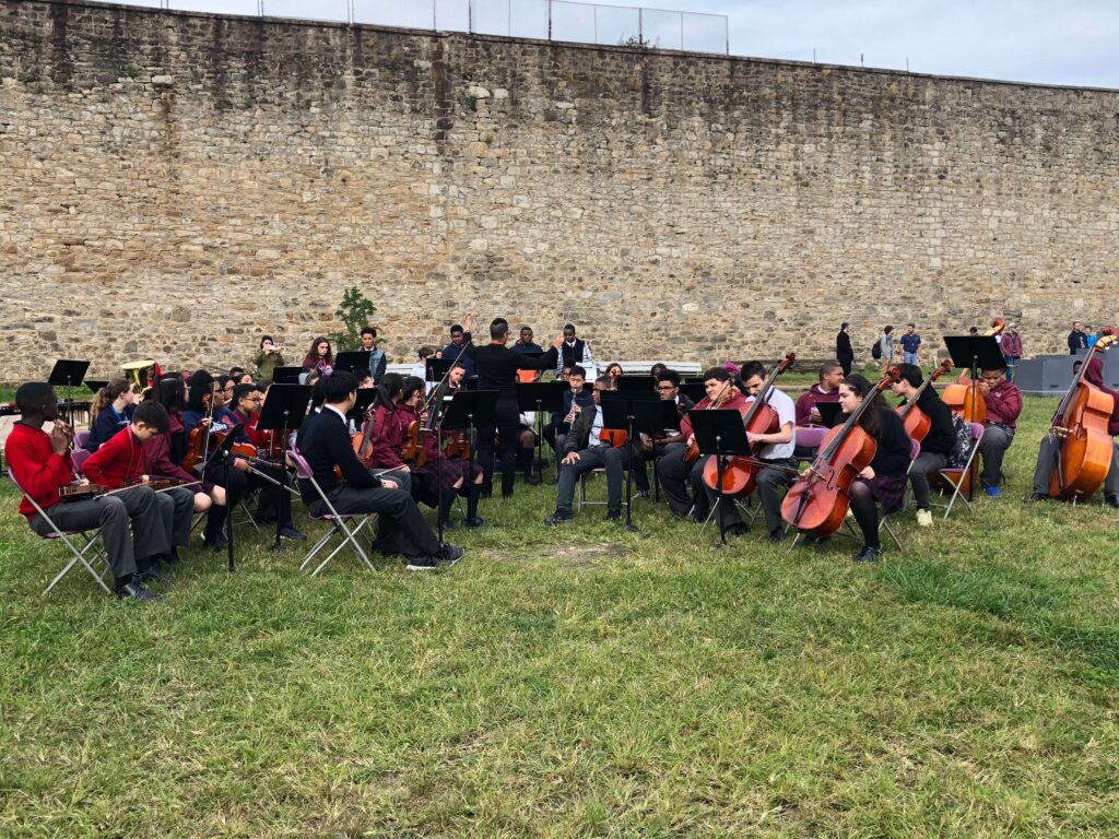 Group of children play orchestral instruments outdoors.