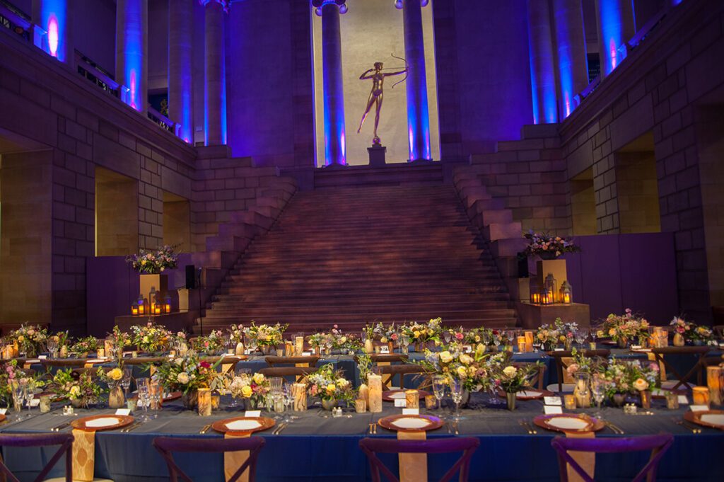 Blue lights illuminate the space, a state stands tall in the middle at the top of a staircase inside of the Philadelphia Museum of Art. Tables, set for an elaborate affair with floral arrangements and candles, are at the bottom of the steps