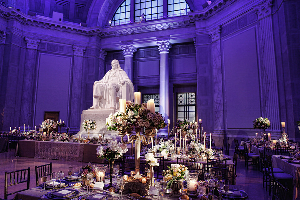 A large sculpture of Benjamin Franklin is in the center of the room, purple lights illuminate the space, beautifully decorated tables with elaborate floral centerpieces surround the statue, with the space ready to host an event as the tables are set for an elegant affair.