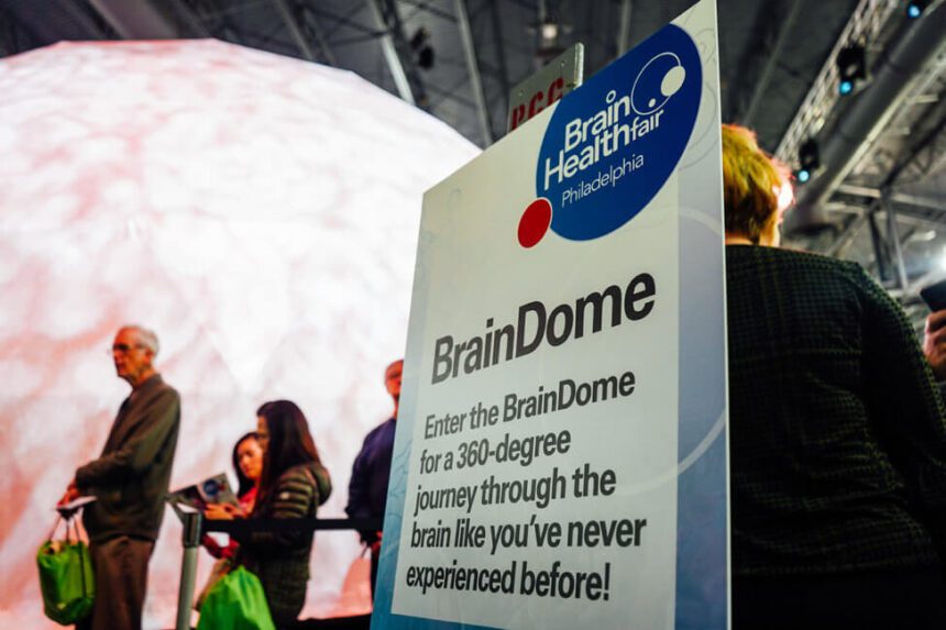 There are people standing in line, waiting to enter a large white dome. In front of them, a large white sign reads Brain Health Fair Philadelphia Brain Dome Enter the Brain Dome for a 2360-degree journey through the brain like you've never experienced before!