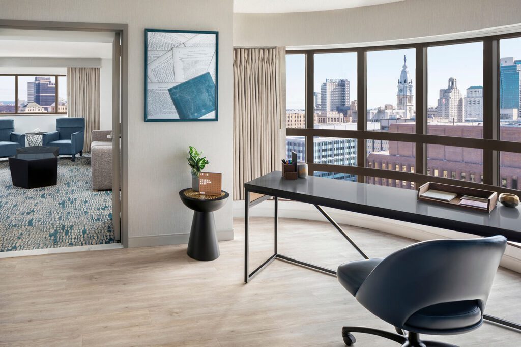A desk chair on wheels is off to the right in front of a desk, both facing a large window showing the city's skyline. Through a doorway to the left, there is a sitting area with two light blue chairs and a small table in front of them.