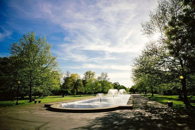 A bright sky is overhead with wispy clouds. Trees line a cement path, in the middle is a large fountain with three sprinklers. Benches also line the pathway.
