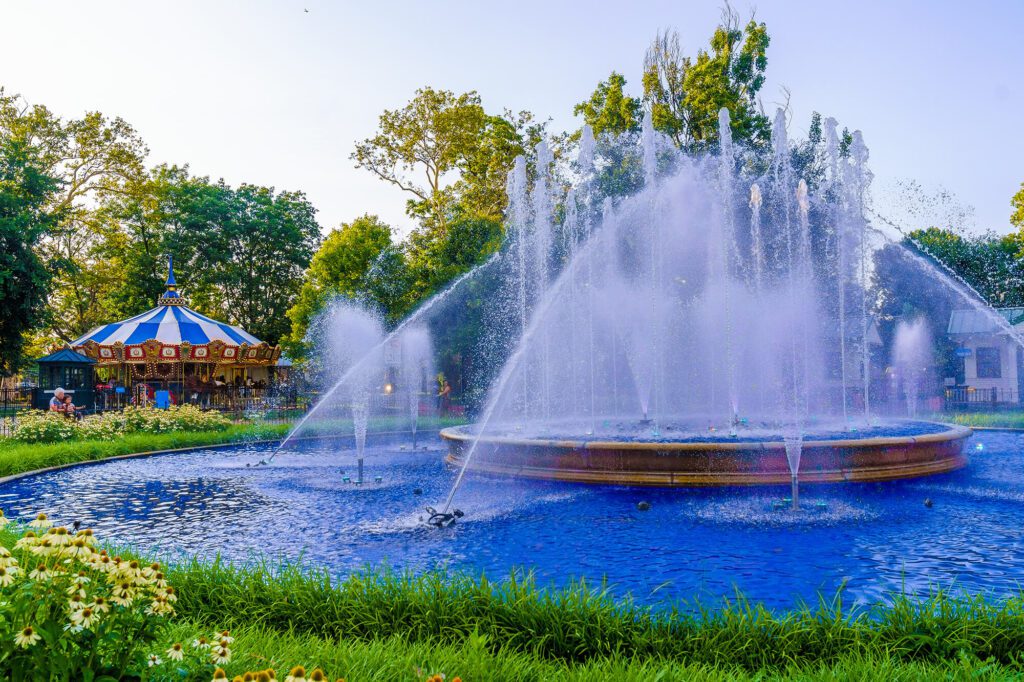A carousel if off in the distance to the left. A large fountain comes to life in the center, with green grass surrounding it, along with yellow flowers. Trees are in the background under a clear blue sky.
