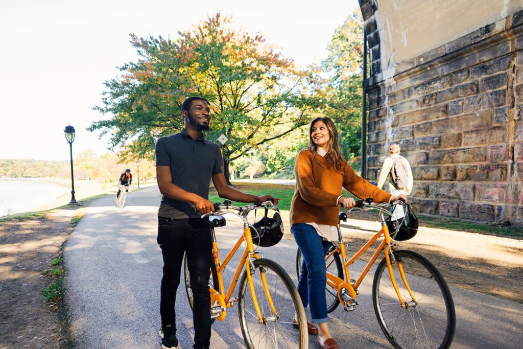 Two people stand next to bikes. A man is off to the left. A woman stands to the right. Both bikes are yellow and black. a man walks behind them in the opposite direction as an oncoming bicyclist heads toward them. Trees are in the distance.