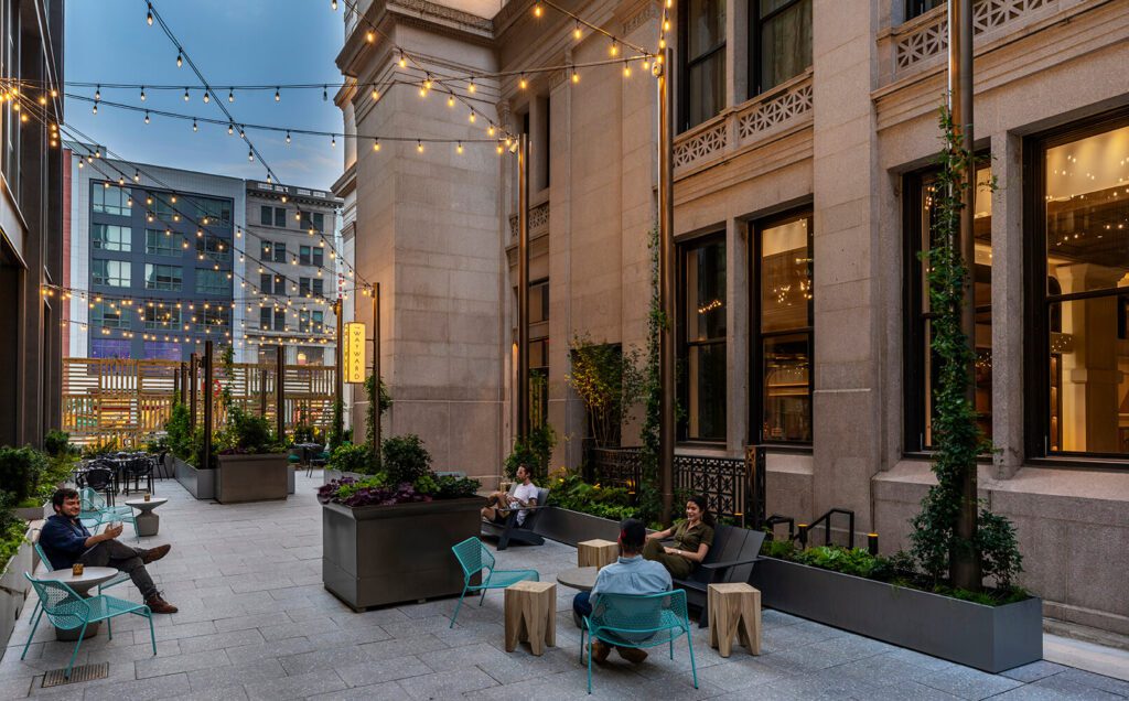 Stands of lights hang overhead as four people sit on blue chairs spread throughout the outdoor space decorated with large square flower pots with greenery