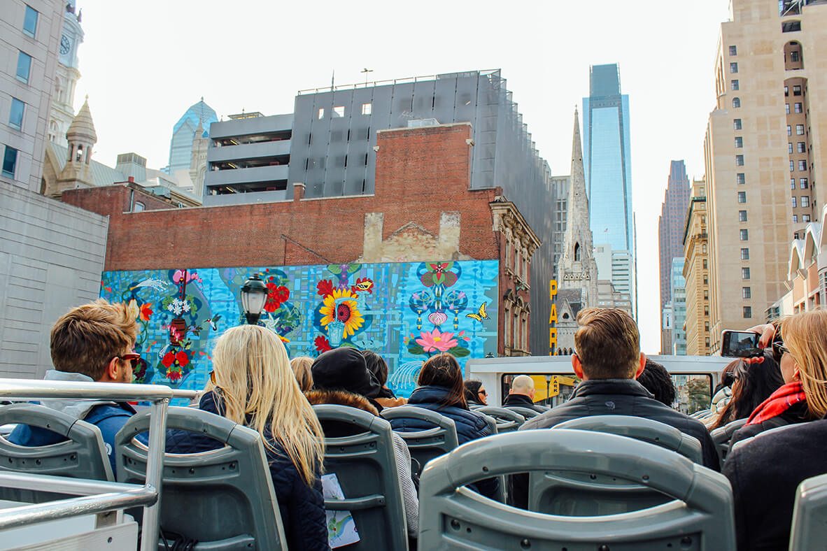 A group of people are shown sitting on top of a tour bus. A bright blue mural is to their left. Skylines are in front of them.