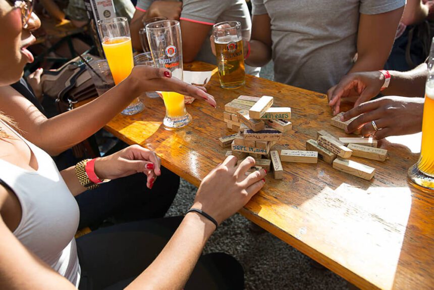 Hands are shown playing Jenga on a picnic table. Beers are shown on the table.