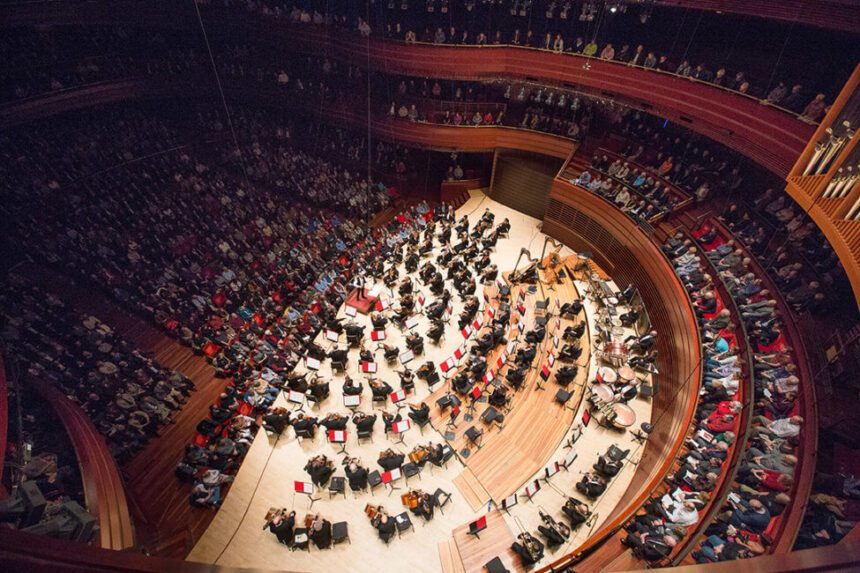 An overhead view on the orchestra on stage. Audience members are seen in the seats both in front and behind the performers.