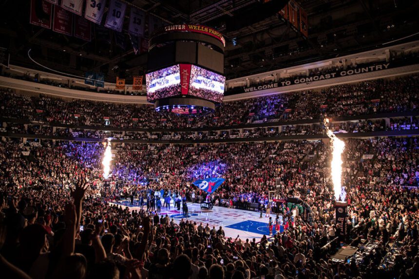 A packed indoor arena is shown. Fans fill the stands. Every seat is filled. Fire shoots from either end of the basketball court. Players are shown on the court. A large blue flag with a red 7 and a white 6 is shown in the center. The jumbo tron hanging above the court lights up.