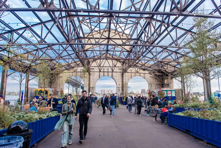 An open-air market is underway at Cherry Street Pier. A couple is shown walking off to the left, but toward the camera. Greenery is spread throughout the spaces. Some people are shown walking around, others are shown sitting around tables. The river can be seen in the distance.
