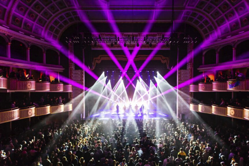 Purple and white lights light up the stage inside of an indoor music venue called The Met. Audience members are shown standing facing the stage.