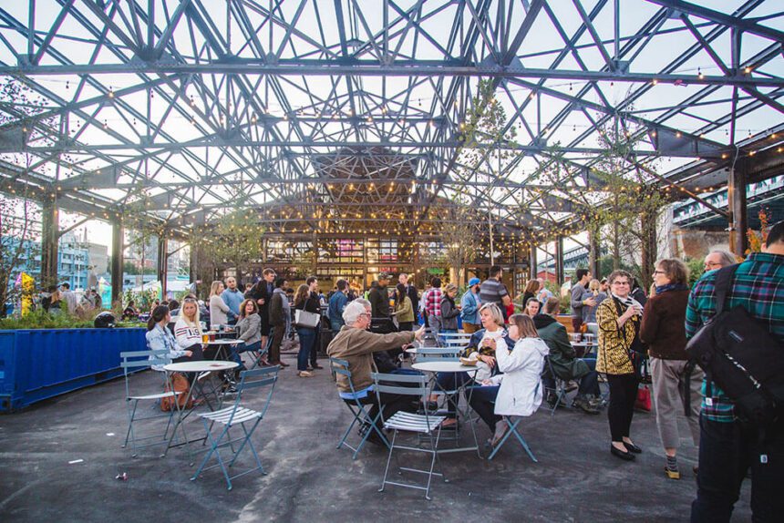 An open-air pier with an open roof is set up with metal chairs and tables. People are shown standing around the space. Others are sitting down. Lights are strung overhead. Greenery is also spread throughout the space.