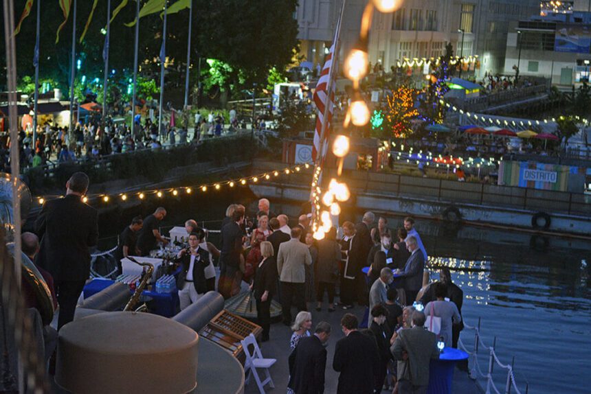 It is nighttime. Lights are strung overhead. A large group of people are shown on a deck of a ship enjoying the fresh air. More lights are shown above the boardwalk to the left and a barge to the right