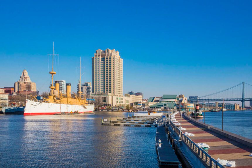 The bright blue sky above is clear of any clouds. A skyscraper is shown in the distance. In front of it to the left is a yellow, white, and red ship. A walkway is shown off to the right above the water below it. A bridge is shown off in the distance to the right.