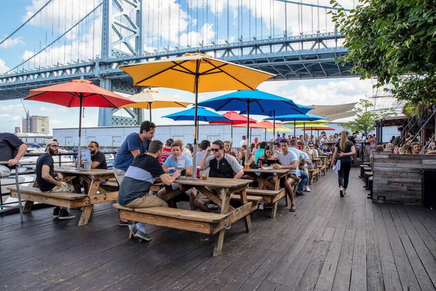 The Benjamin Franklin Bridge soars overhead beneath a blue sky with white fluffy clouds. Picnic tables are set up on the pier. Yellow, red, blue, orange, and yellow umbrellas provide shade for each table. People are shown sitting at the tables. A server is off to the right walking toward customers.