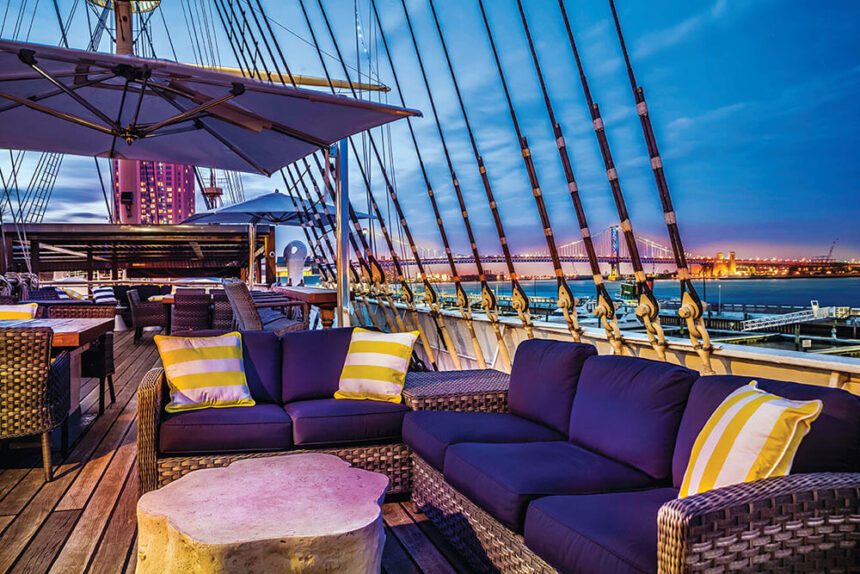 An outdoor sofa is shown on the deck of the Moshulu, a historic ship. The sofa has dark blue cushions. There are three yellow and white striped pillows on the couches. Off in the distance is a bridge. The sky above is a bright blue. 