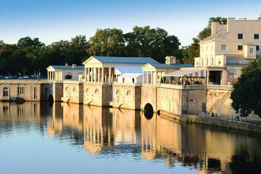 A clear blue sky is overhead. Trees are shown in the background. The focal point is a large white structure up against and reflecting into the river below. The water is calm. A balcony toward the right appears to be hosting an event with people.