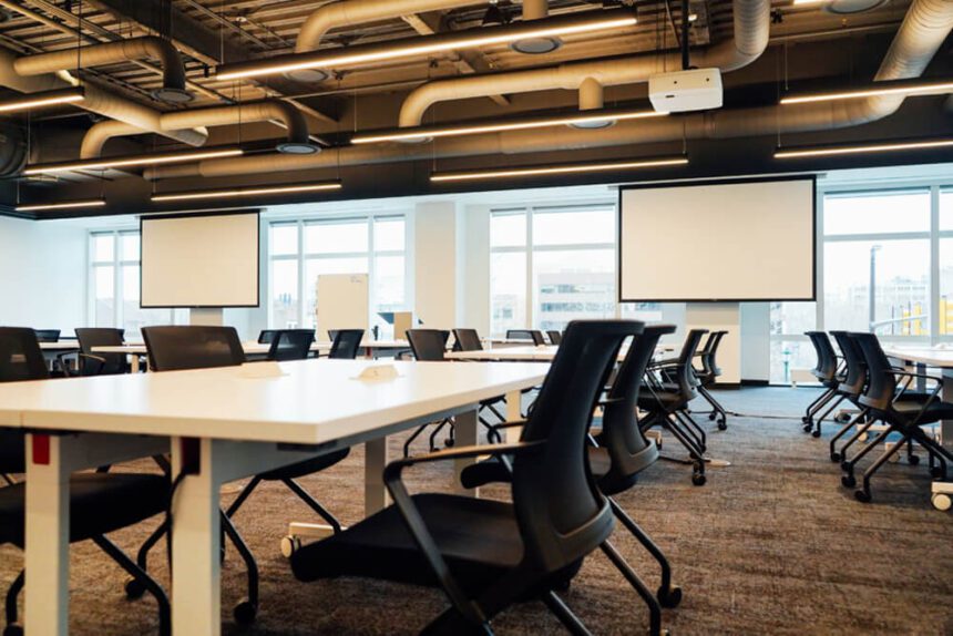 White desks are shown with black shares pushed up against them. Two large white screens are shown against windows that are letting in natural light.