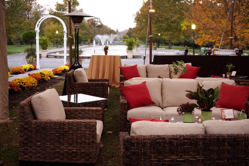 An outdoor patio set is set up. The chairs and sofas are brown wicker with beige cushions and red-orange pillows. Tables in front of the sofas are set with planters and candles. Behind the sofas, there is a high top cocktail table covered in a beige tablecloth. A flower is placed in the center. Beyond that, there are fountains spraying water into the air. Fall foliage is seen on either side of the fountain pool. Yellow and orange mums line a walkway to the left of the seating area.