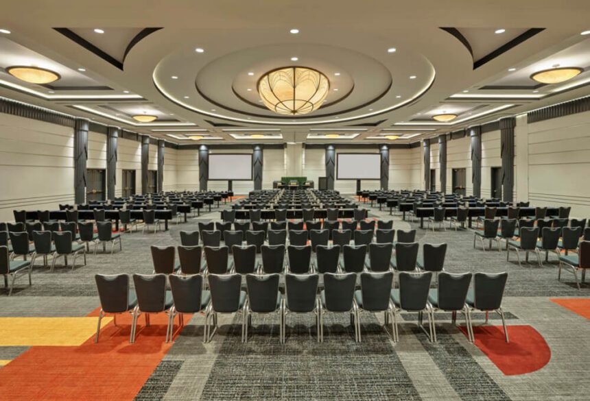 A large rom is shown set up with rows of gray cushioned chairs. The chairs are facing a desk set up in the center. Two large screens are shown, one on either side of the main table up front. The carpet is a mix of gray, yellow, and orange designs.