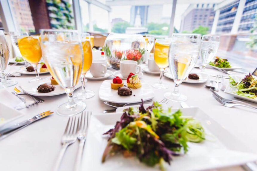 A spread of plates of food and drinks are set up across a table covered with a white tablecloth. The plate in the center has a salad made of green and purple leaves. A plate in front of the salad has three small desserts on it. There is one that appears to be chocolate, one that looks like a tart with three raspberries on top, and another pastry of sorts with a whipped portion and a piece of fruit as garnish. Glasses on the table are filled with water and a yellow juice. A window in the background is letting in natural light. Independence Hall is seen off in the distance.