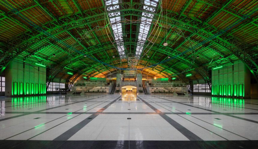 A massive hall is shown lit up in green. The floors are a mix of white and gray. They shine reflecting the lights above. Hanging from the industrial roof above is a sculpture made of blue and silver metals. A hallway at the end of the hall lights up leading to conference rooms.