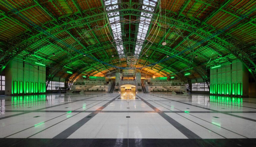 A massive hall is shown lit up in green. The floors are a mix of white and gray. They shine reflecting the lights above. Hanging from the industrial roof above is a sculpture made of blue and silver metals. A hallway at the end of the hall lights up leading to conference rooms.