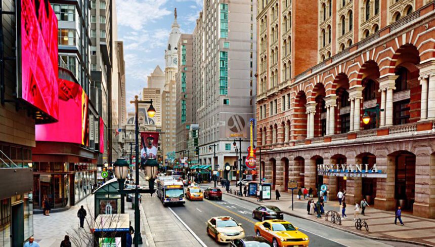 To the right is the Pennsylvania Convention Center. In front of the center is a street filled with cars, taxis, and a SEPTA bus. There are people walking along the sidewalk in between the center and the street. The Marriott hotel is shown next to the center. Philadelphia City Hall is in the background.