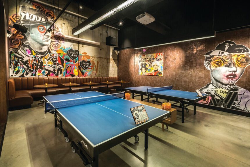 Two blue and white Ping-Pong tables are shown in the middle of a room. The far back wall has a colorful mural painted on it. Underneath the mural, the wall is lined with cushioned booths with five small tables in front. On the wall to the right, another colorful mural decorates the space.