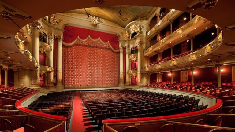 A large theater is shown. There are rows of red seats. The stage is front and center. There are gold accents and pillars throughout the space.