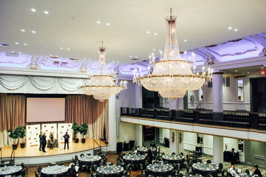 There are two very large chandeliers hanging from the ceiling. Underneath, there are several tables set up with black tablecloths and place settings at each chair. There are four people shown standing on a stage off to the left. There are also four large plants on the stage. The trees on either side of the backdrop. A small table is in the center of the stage.