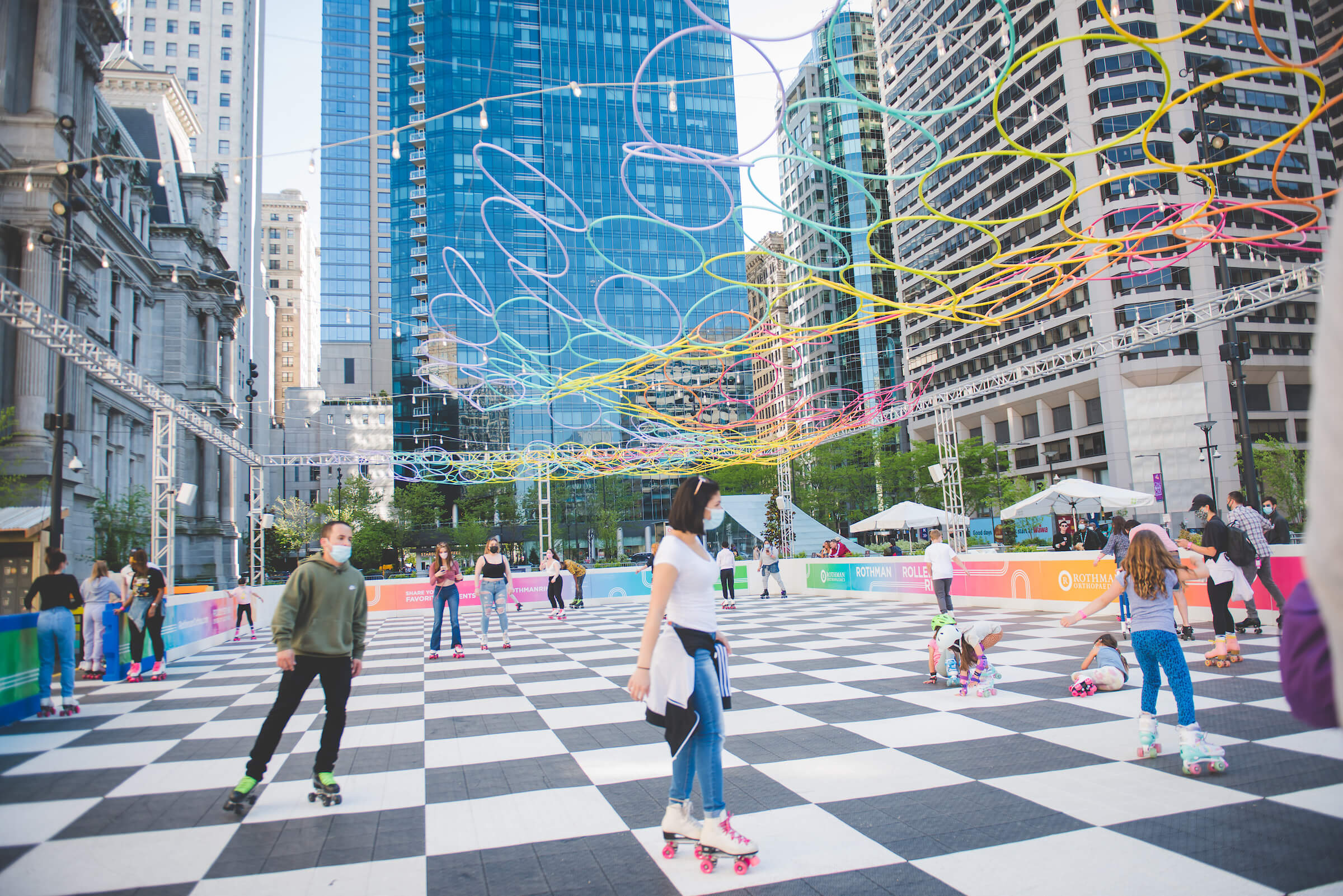 People roller skating on a checkerboard rink in the city as skyscrapers tower over them.