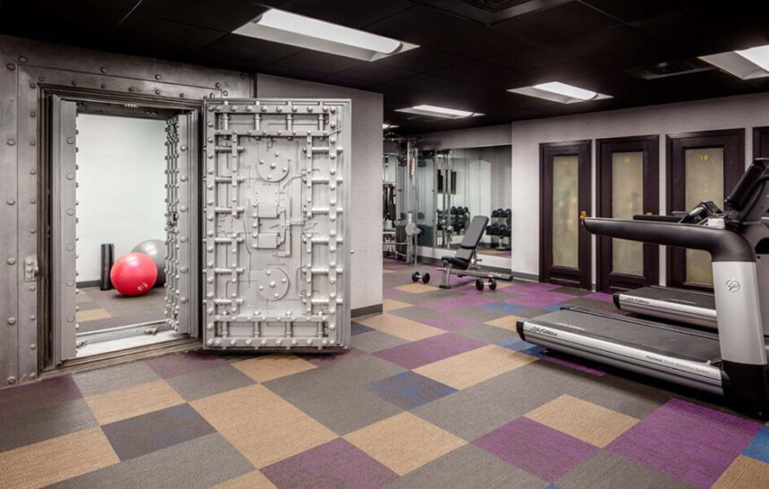 An indoor fitness center is shown. A large red yoga ball is shown in a room off to the left. It appears to be in a room with a vault door. The carpet is gray and purple. There are two treadmills off to the right. Beyond that, there is a space with weights.