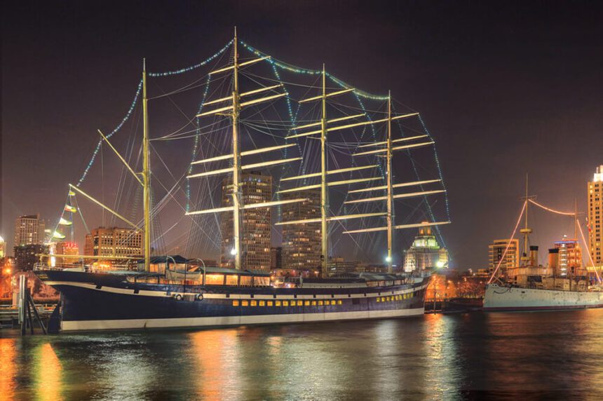 It is night time. The sky is dark. A large ship is shown on the water. There are bright blue lights hung outlining the ship. The river below reflects the bright lights. Another smaller ship is nearby, also docked along the waterfront.