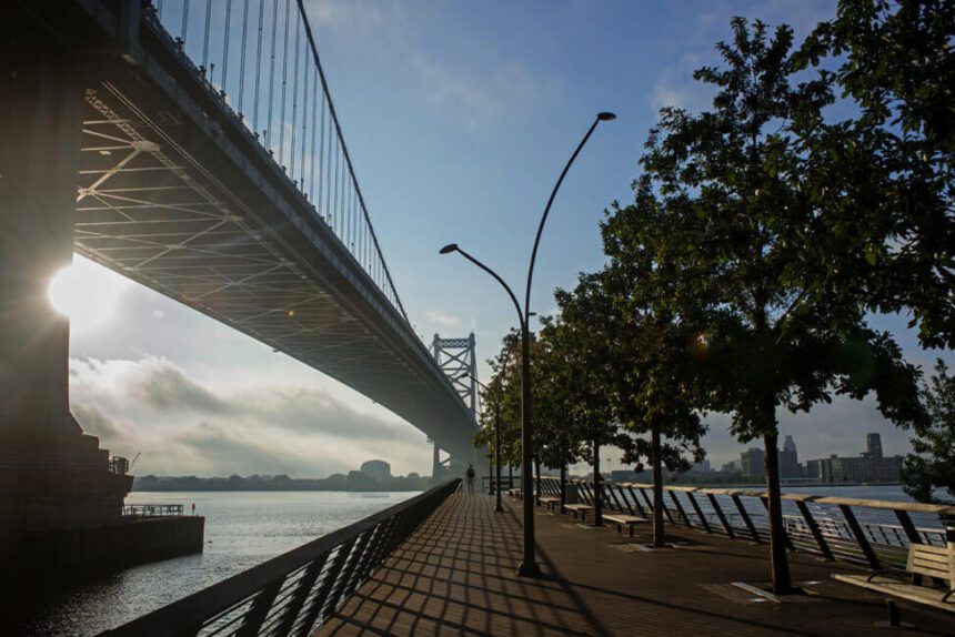 A large bridge soars overhead on the left. A pier stretches down the middle. To the right, there are trees and benches lining the pier. The river below reflects the sunlight coming from above.