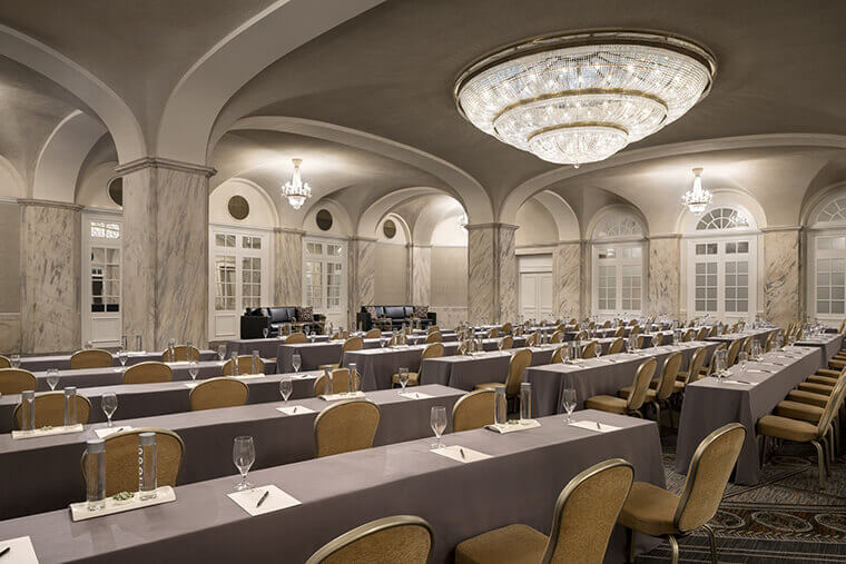 There are yellow and gold chairs pushed up against rows of gray tables spread throughout a meeting room. There is a large chandelier hanging from the ceiling in the middle of the room. Large marble pillars are shown. Beyond them are two large black couches with decorative pillows.