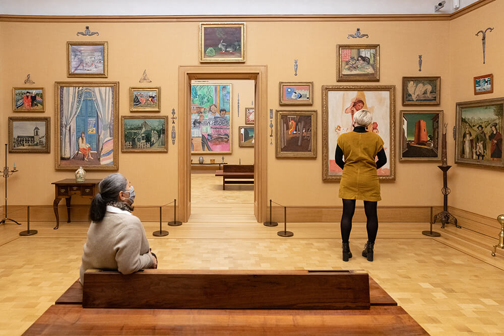 A woman is shown sitting on a bench to the left, while another woman is shown standing off to the right. They are both in a room surrounded by art, admiring the framed, colorful works hanging on the walls around them.