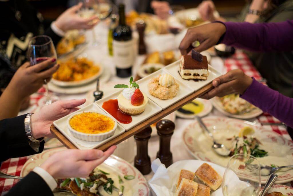 A table full of plates of food is in the center. Two people are passing a white rectangular plate full of different desserts