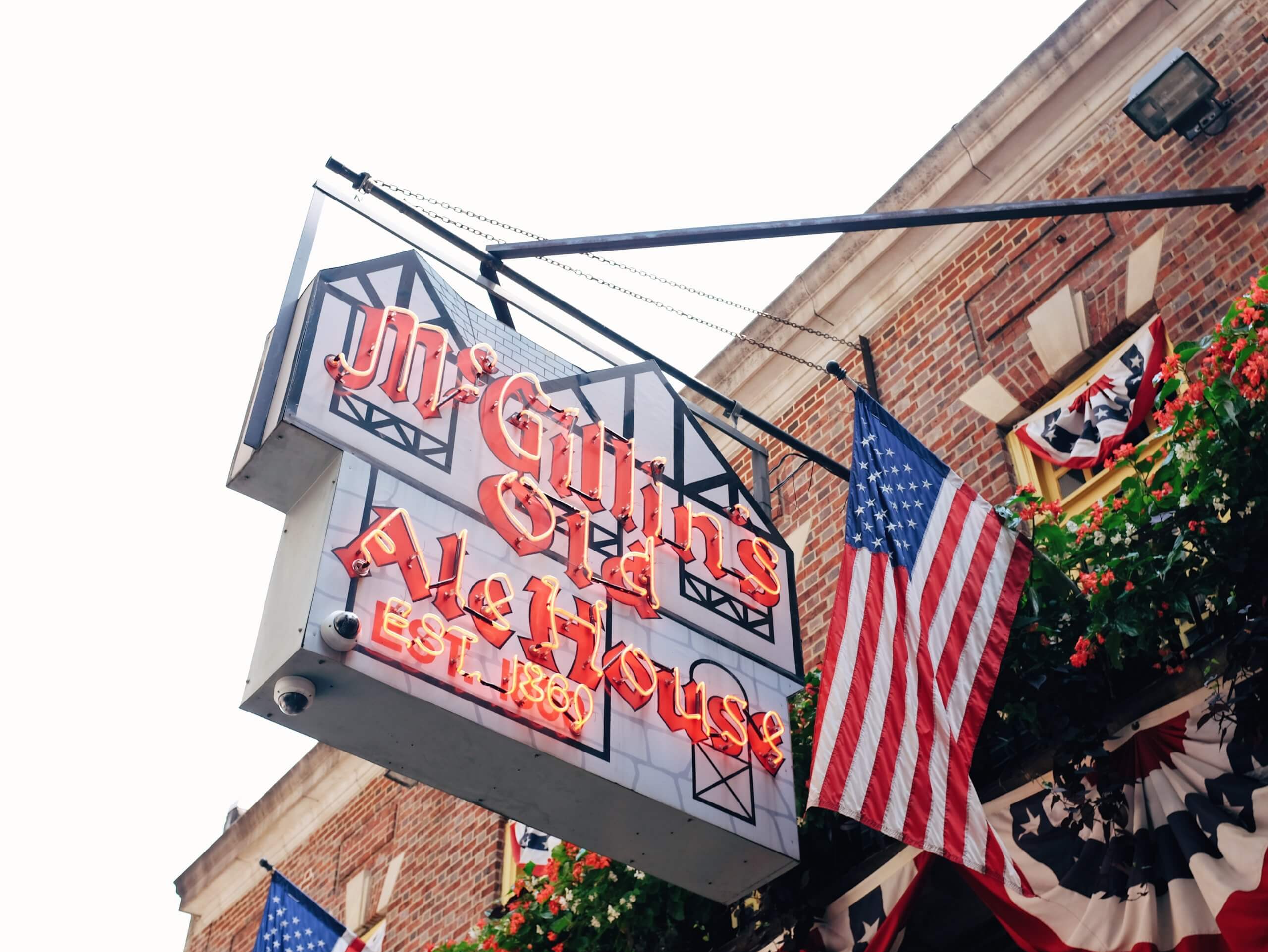 A neon sign for McGillin's Old Ale House hangs outside of the establishment. An American flag is hanging beside it. Both are attached to an old, brick building on the right