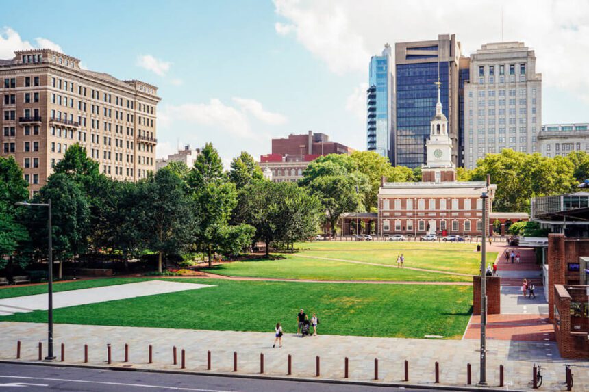Independence Mall is shown. A large lawn of lush green grass is in front of Independence hall. There are people shown standing on the sidewalk taking a picture. The sky above is a light blue with fluffy clouds. There are dark and light green trees lining the outdoor space. There are skyscrapers beyond the historical building.