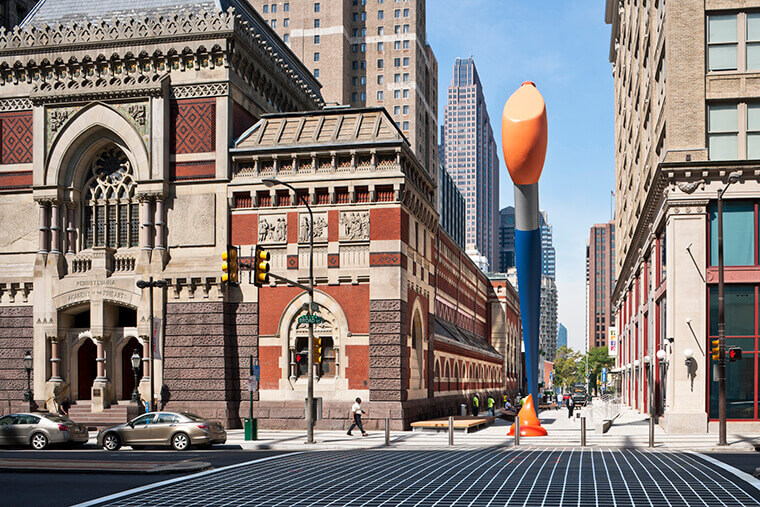 A large sculpture of a paintbrush is shown. The paint is orange, the handle of the paintbrush is blue and silver. There appears to be a glob of orange paint on the sidewalk. The sculpture is shown in between buildings. A man walks across the sidewalk below it.