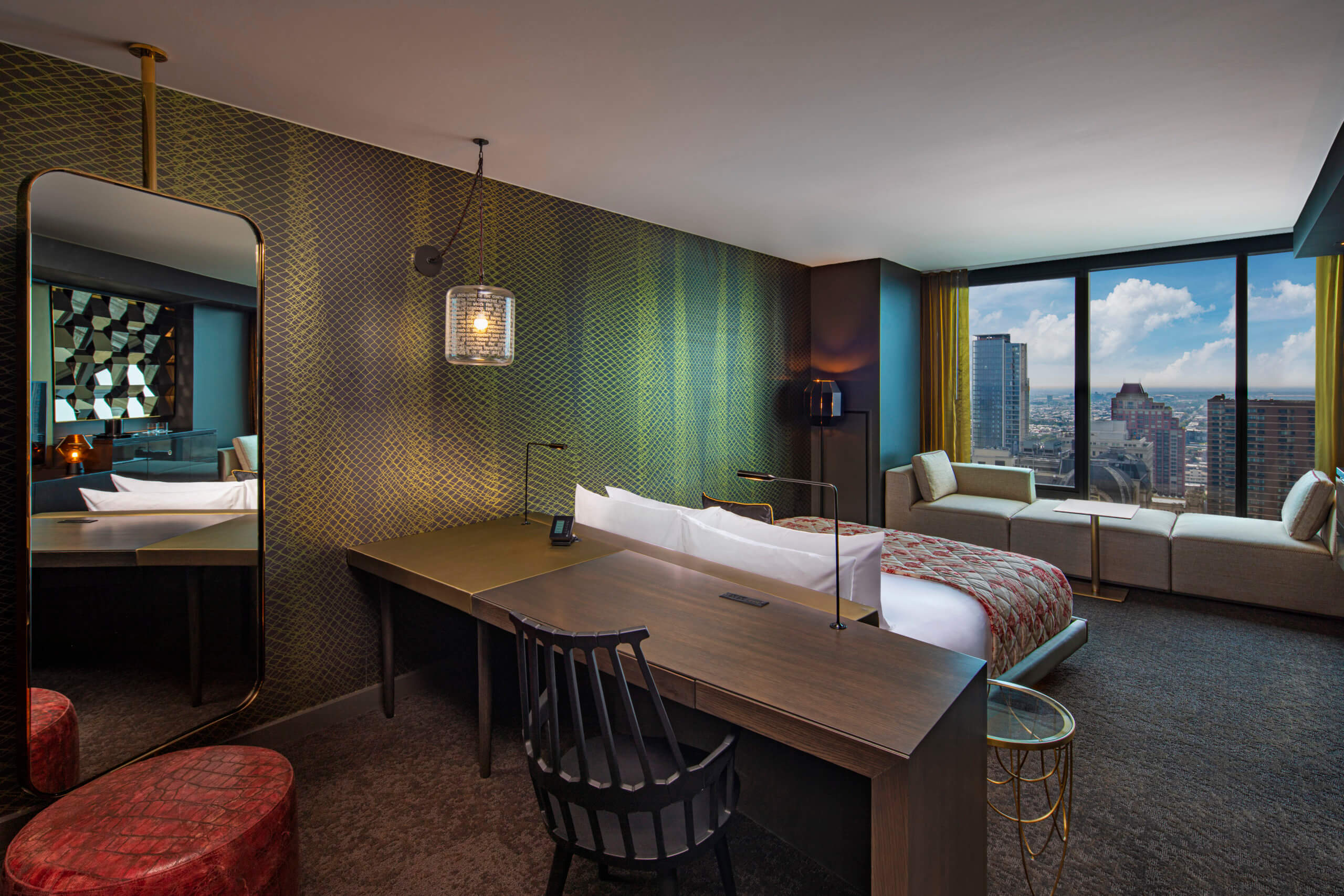 A desk sits behind a bed that faces a window, a green wall is to the left of the desk and bed inside of the hotel room