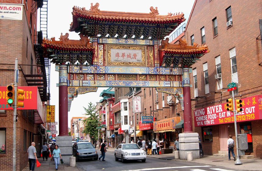 A colorful archway called the Friendship Gate leads to Philadelphia's Chinatown. Cars are shown in the street, as people walk along the sidewalk across colorful stroefronts.