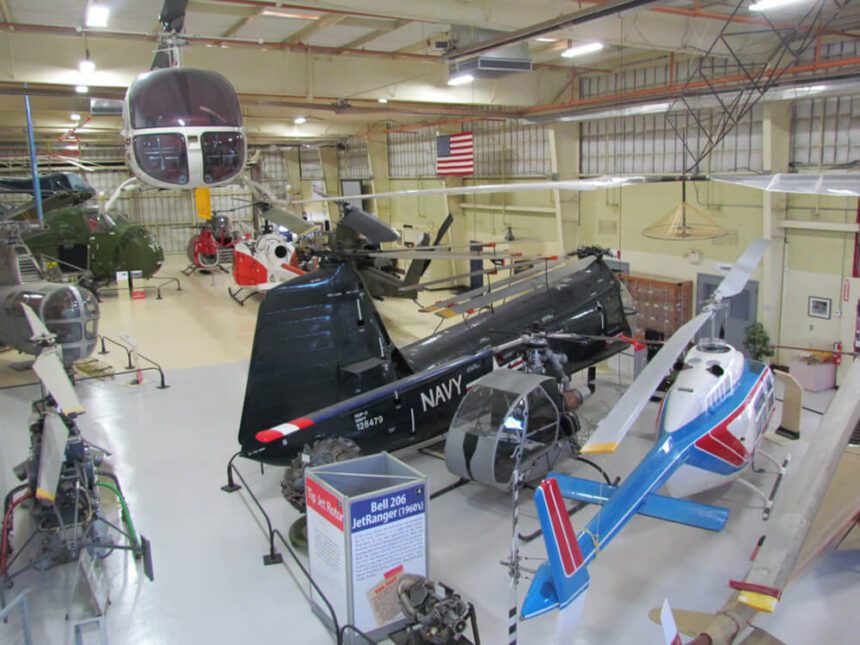Helicopters are set up inside of a warehouse. The one on the right is red, white, and blue. The one next to it, to the left, is navy blue with the word Navy written across it in white. In the background there are multiple helicopters. An American flag is hanging up against a wall.