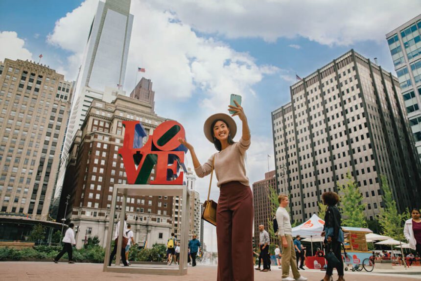 A woman wearing auburn pants, a beige sweater, and a tan hat poses for a selfie in front of the iconic red LOVE sculpture in LOVE Park. People throughout the space are shown behind her.
