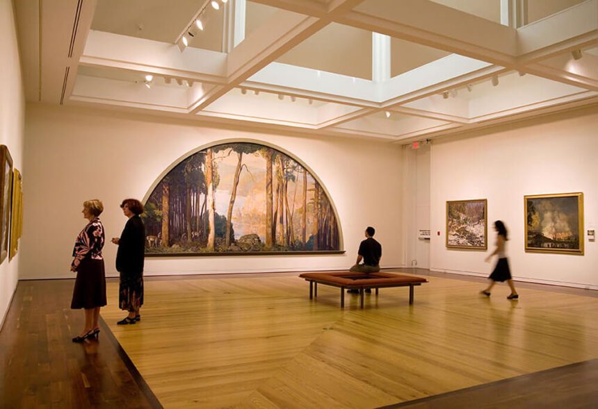 A large exhibit space is shown. Two women are standing admiring work on the wall off to the left. A man is shown sitting on a bench in the middle of the room as he admires a large work of art hanging on the wall in front of him. A woman appears walking off to the right. Two other works are hanging on the wall beside her.